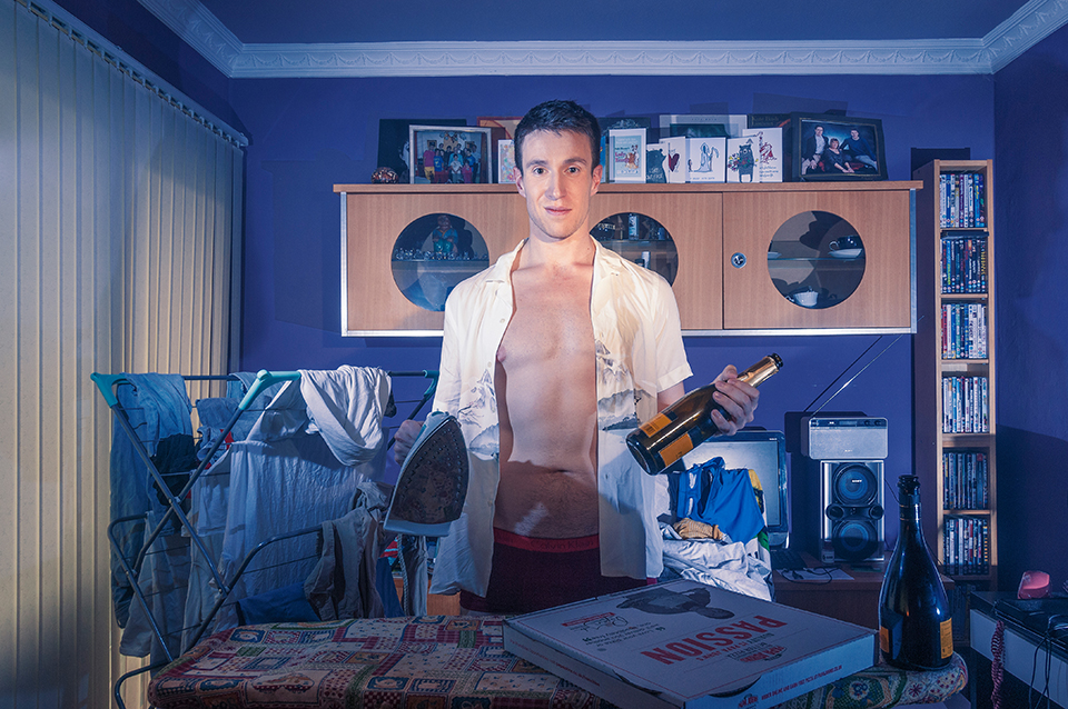 Male subject in messy flat with laundry, pizza box and bottle of Prosecco