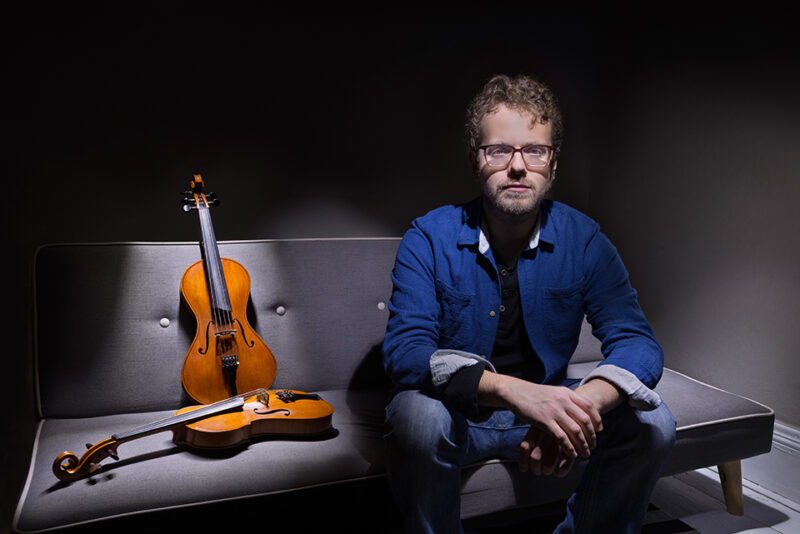 Musician Tom Fairbairn sitting on a couch with two violins